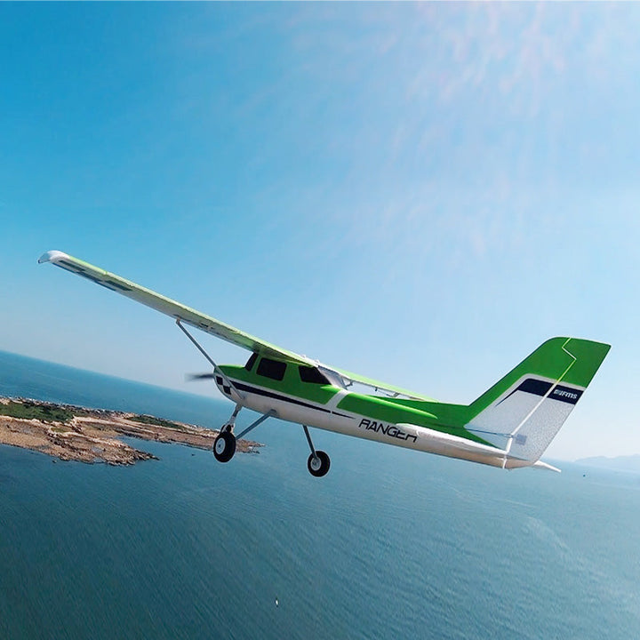 Green and white remote control model airplane flying over the ocean with coastline and mountains in the background under a clear blue sky.