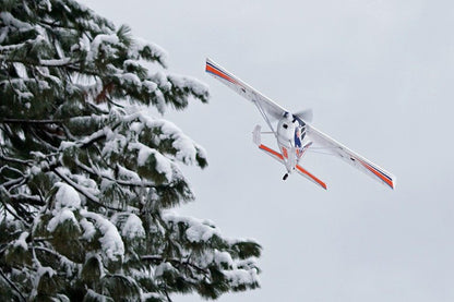 FMS 1400mm Kingfisher PNP aircraft in flight, banking left near snow-covered pine trees.