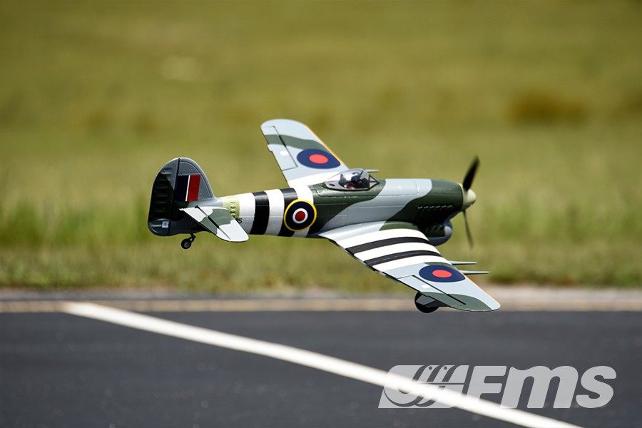 FMS 1100mm Typhoon PNP model airplane in flight, featuring camouflage paint and RAF roundels.